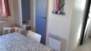 Ein schlichter Raum mit einem Bett, einem Tisch mit gemustertem Tischläufer, einem Fenster mit Vorhängen, einem Wandheizkörper und einer Tür.