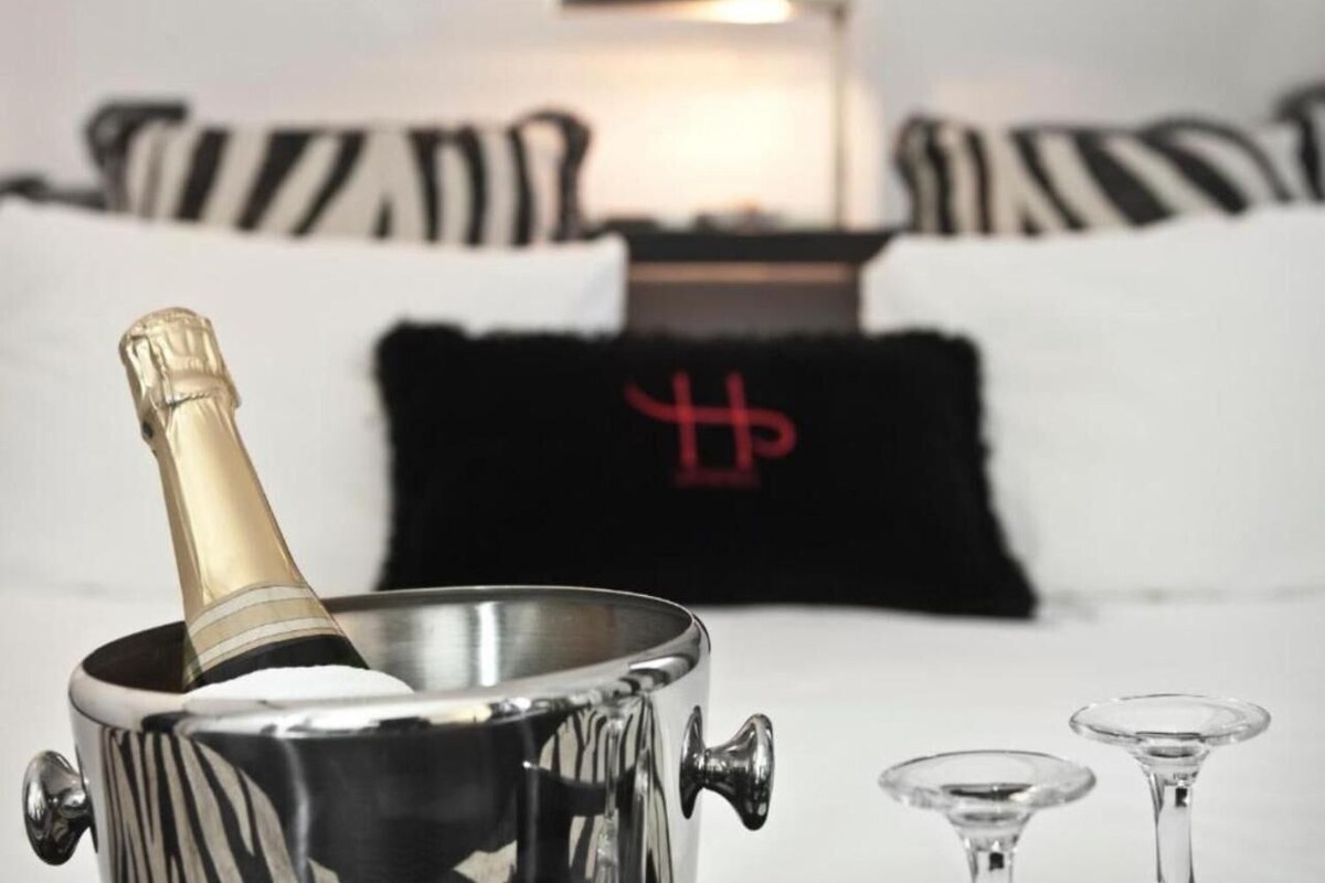 Een metalen champagnekoeler met een fles, twee wijnglazen en een zwart kussen met een rood ontwerp in een moderne kamer met gestreepte beddengoed.