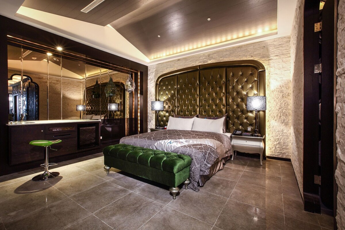Une chambre à coucher luxueuse, dotée d’un grand lit, d’un banc recouvert d’un tissu vert, d’une coiffeuse avec un miroir et d’une table de chevet avec une lampe.