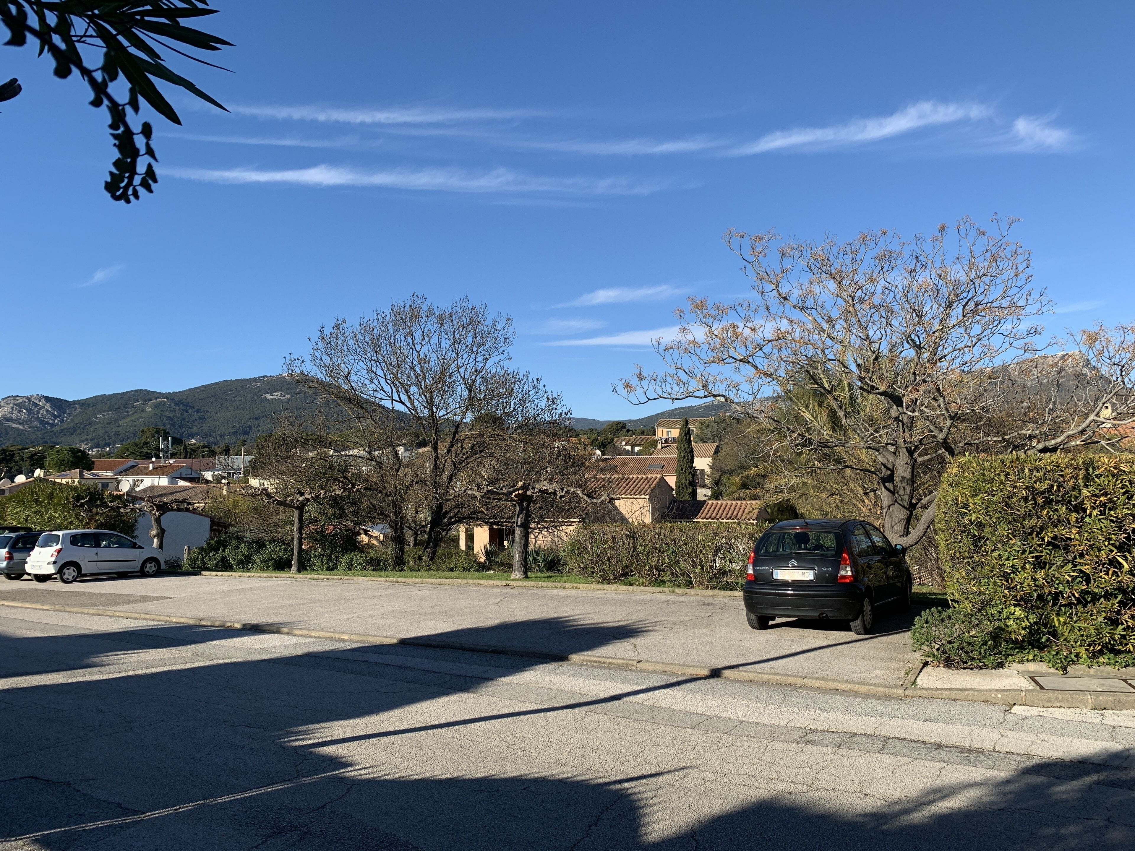 A parking area with a car parked near a building with a tiled roof, surrounded by trees and hills in the background.