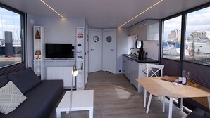 Een moderne woonkamer met een bank, een salontafel en een televisie. Er is een keukenhoek met een eettafel en stoelen. De kamer heeft grote ramen met uitzicht op de jachthaven.
