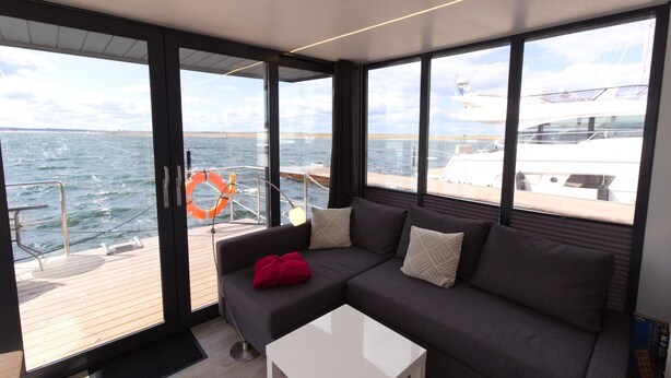 Een modern interieur met een donkergrijze bank, een witte salontafel en grote ramen met uitzicht op zee en jachten.