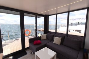 Un interior moderno con un sofá gris oscuro, una mesa de centro blanca y ventanas amplias que ofrecen vistas al mar y a yates.