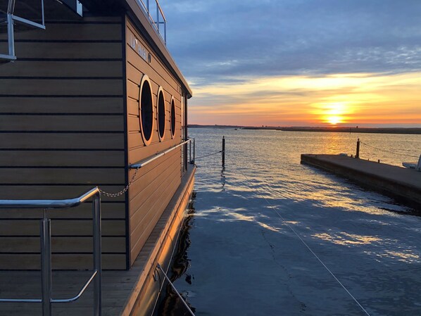 Un edificio de madera con amplias ventanas, un muelle que se adentra en aguas tranquilas y un atardecer al fondo.
