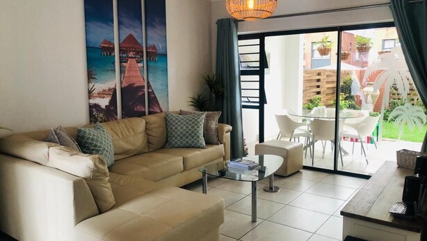 A living room with a beige sofa, a glass coffee table, and a view of a patio with outdoor furniture. - The blyde 3 bedroom crystal Lagoon beach  (Pretoria)