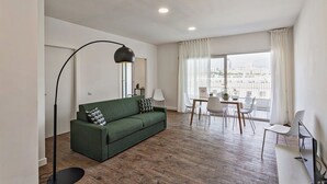 Una sala de estar moderna con un sofá verde, una mesa de comedor con sillas, una lámpara de pie y un amplio ventanal con cortinas.