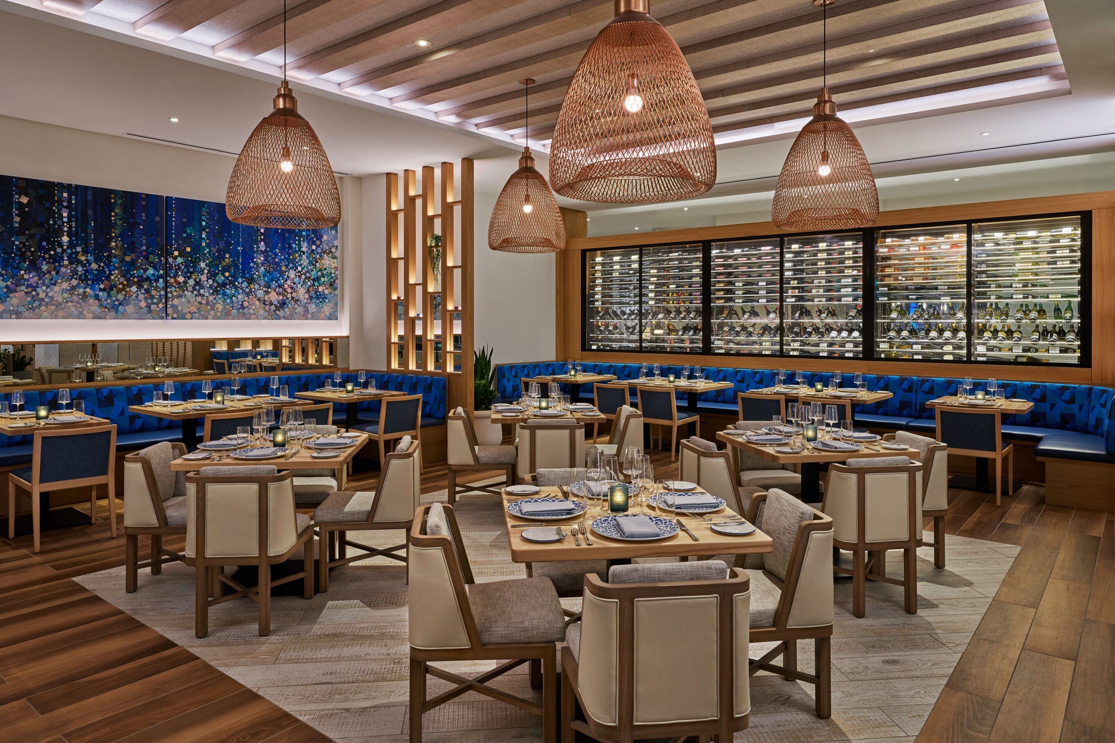 Un restaurante moderno con mesas y sillas de madera, asientos acolchados en tono azul y una amplia exhibición de botellas de vino.