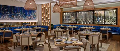 Un restaurante moderno con mesas y sillas de madera, asientos acolchados en tono azul y una amplia exhibición de botellas de vino.