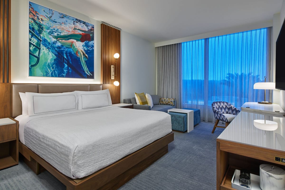 Een hotelkamer met een groot bed, een bureau met lamp, een stoel, een bank en een groot raam met gordijnen.