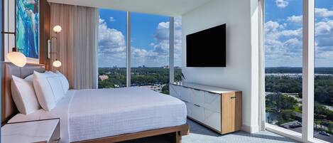Une chambre moderne avec un grand lit, un téléviseur fixé au mur et une vue sur la ville grâce à de grandes fenêtres.