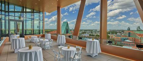 Un amplio espacio exterior con comedor, mesas y sillas redondas, ventanas grandes con vista a la ciudad y techo de madera.