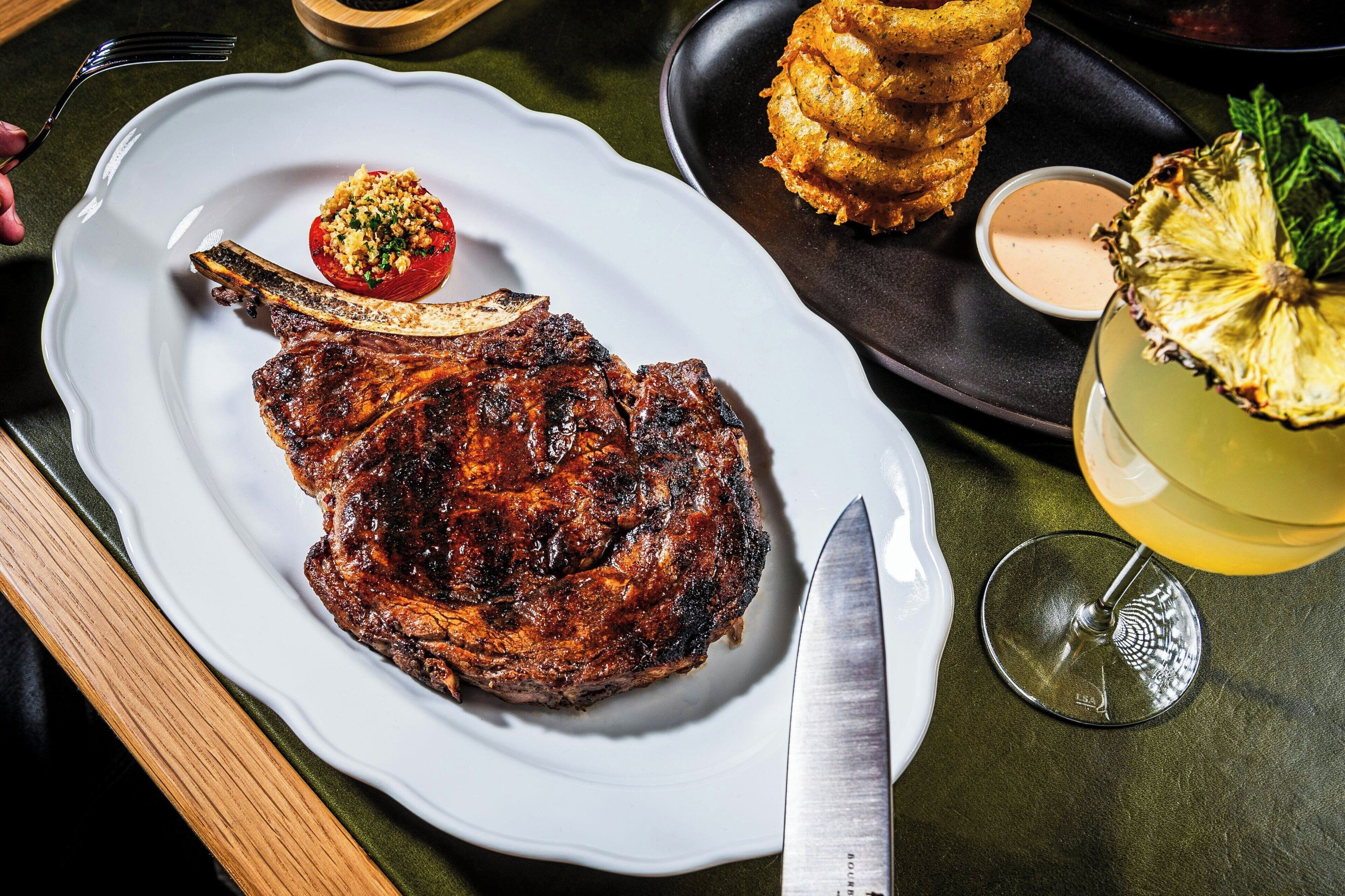 Een bord met een gegrilde steak, gevulde tomaat en gebakken plantains op een tafel.