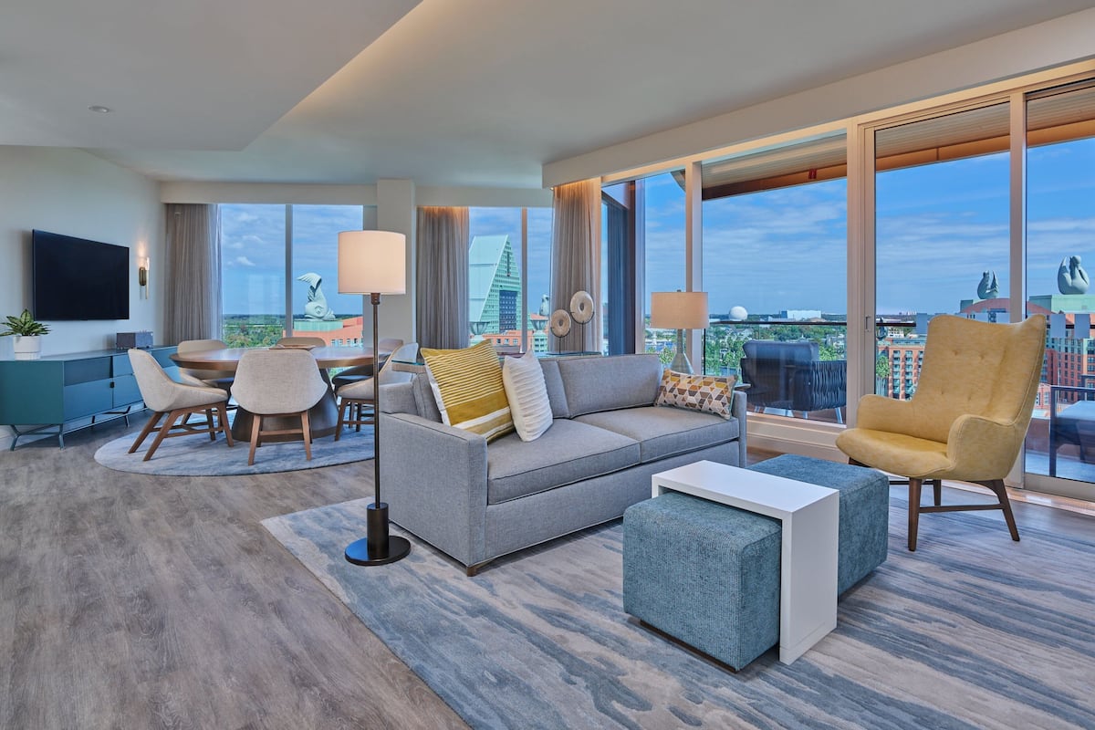 Een moderne hotelkamer met een grijze bank, een gele fauteuil, een eettafel met stoelen, een flatscreen televisie en grote ramen met uitzicht op de stad.