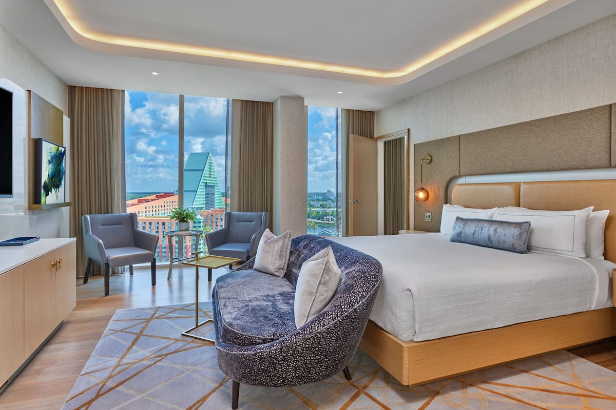 Een moderne hotelkamer met een groot bed, een zithoek met een stoel en een klein tafeltje, een televisie en uitzicht op een stadsgezicht door grote ramen.