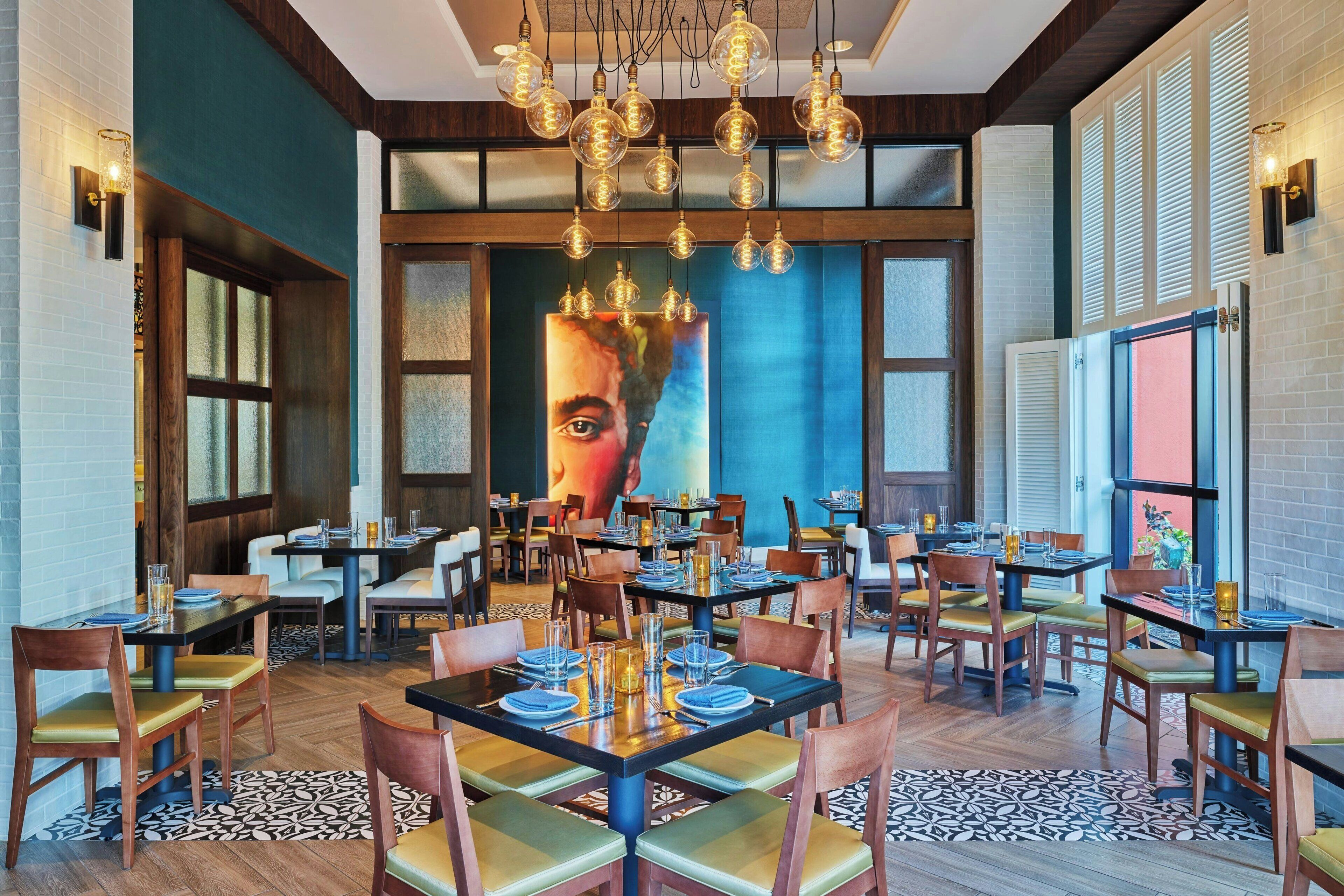 Un restaurante con varias mesas y sillas, un mural grande en la pared y lámparas colgantes.