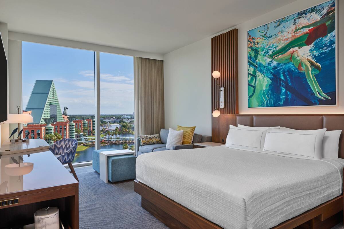 Hotelkamer met een groot bed, een bureau met lamp, een stoel, uitzicht op een stadsbeeld en een schilderij aan de muur.