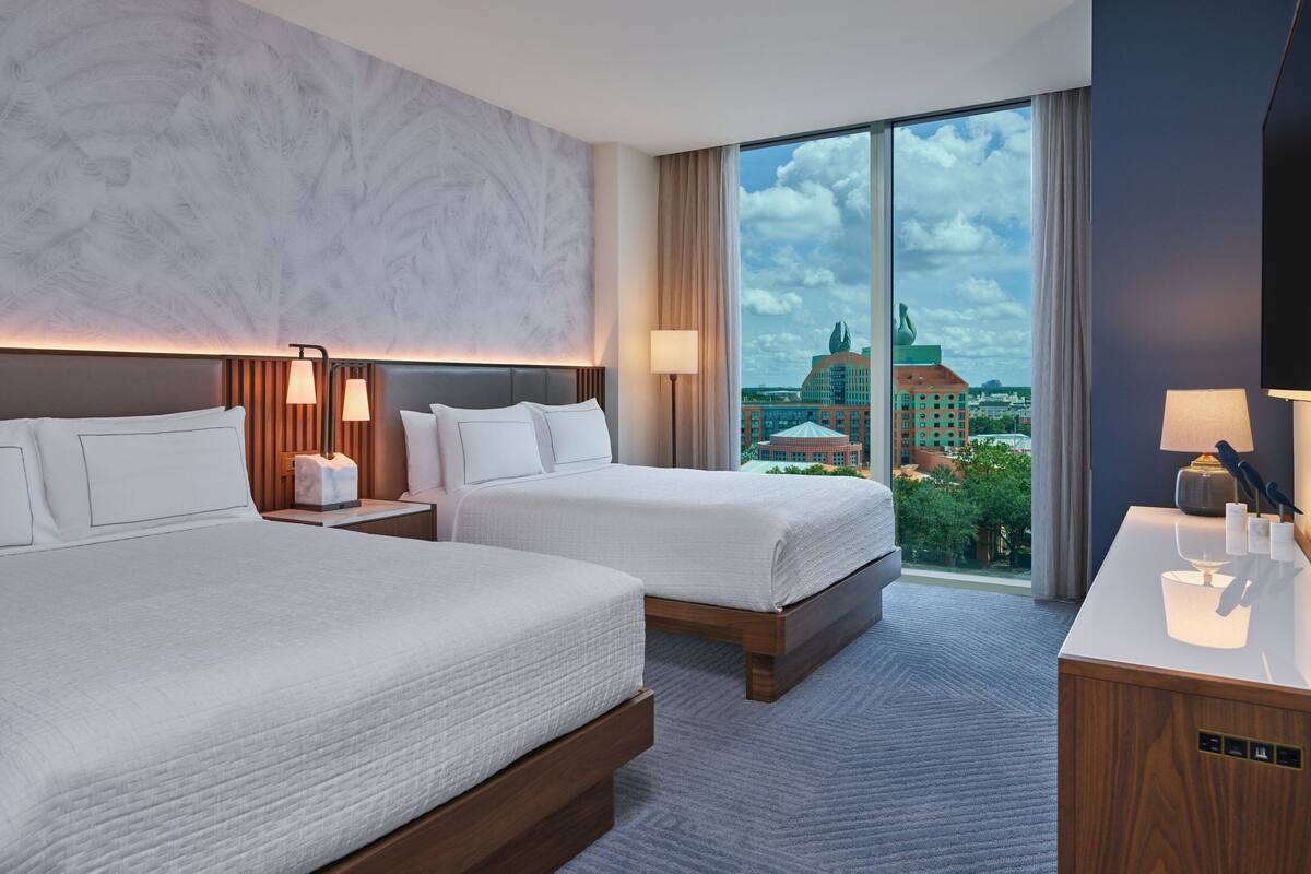 Een hotelkamer met twee bedden, een groot raam met uitzicht op de stad, een bureau met lamp en een wand met een gestructureerd ontwerp.