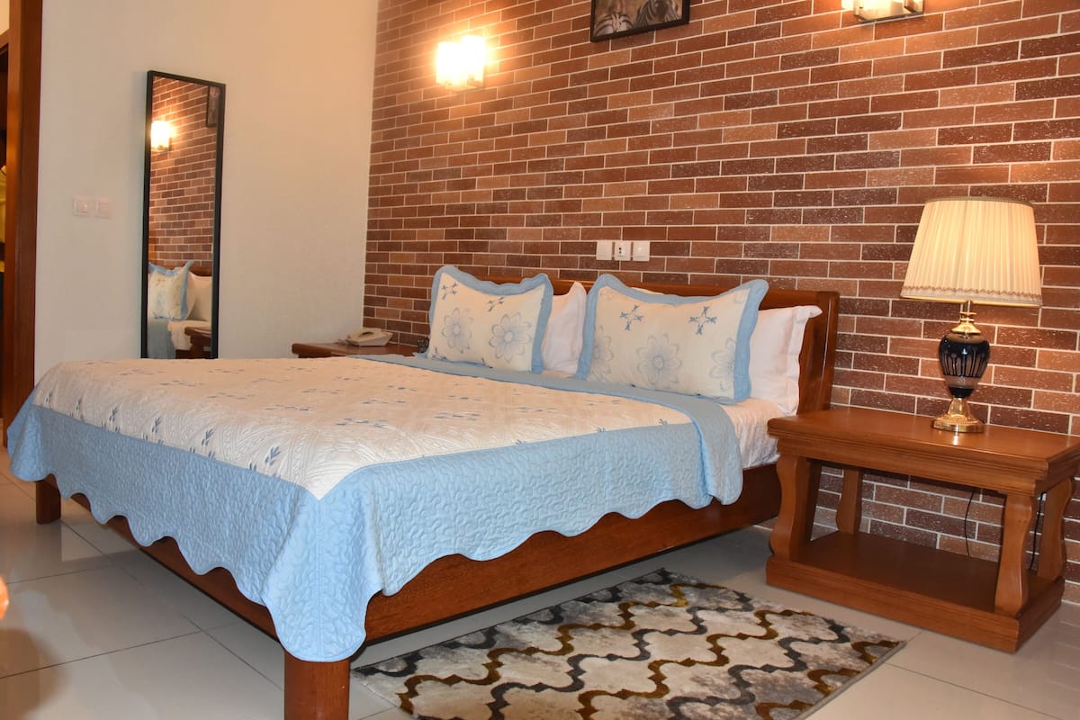 Un dormitorio con pared de ladrillo, cama de madera con colcha azul, mesita de noche con lámpara y alfombra estampada.