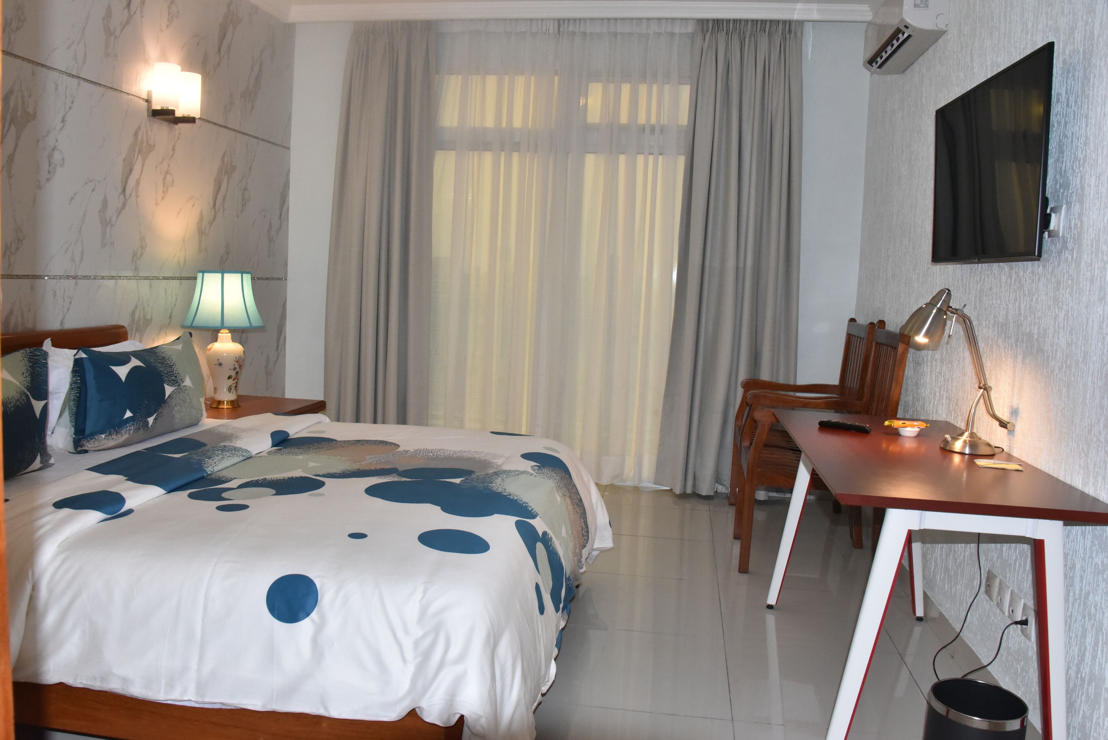 A hotel room with a bed, a desk, a chair, a lamp, and a TV.