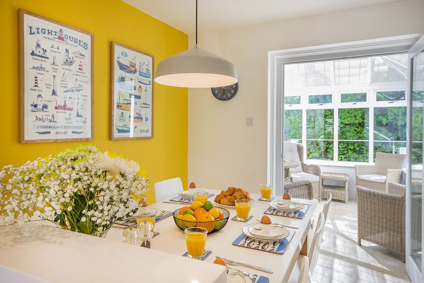 A bright dining area with a yellow wall, a large floral arrangement, and a table set for breakfast with croissants, fruit, and orange juice.