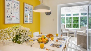 Une salle à manger lumineuse avec un mur jaune, un grand arrangement floral et une table dressée pour le petit-déjeuner, avec des croissants, des fruits et du jus d’orange.