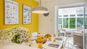 A bright dining area with a yellow wall, a large floral arrangement, and a table set for breakfast with croissants, fruit, and orange juice.