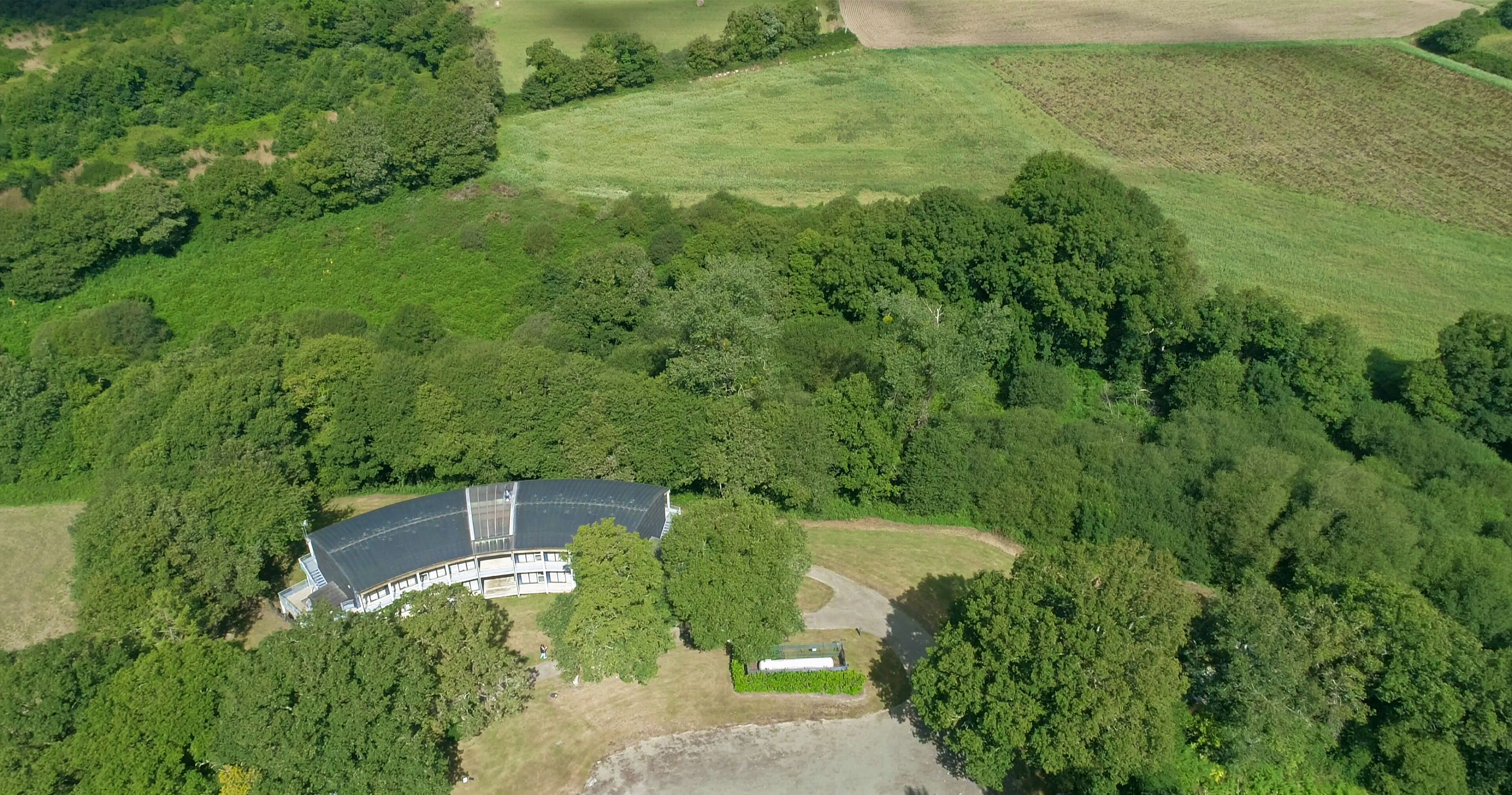 An aerial view of a building surrounded by dense forest, with a large open field to the right.