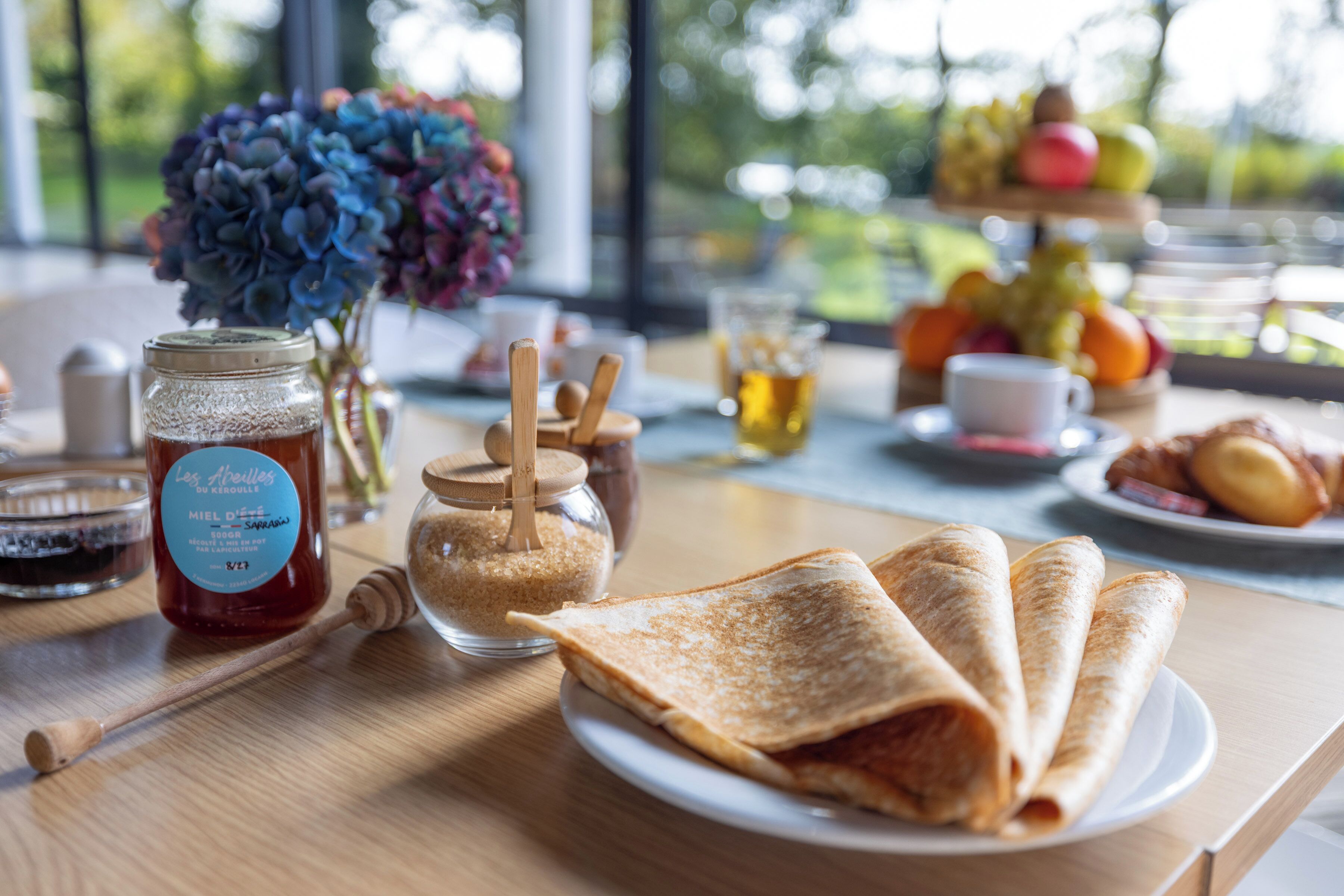 A breakfast table with a plate of crepes, a jar of honey, a honey dipper, a glass of tea, and a vase of flowers.