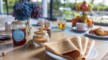 A breakfast table with a plate of crepes, a jar of honey, a honey dipper, a glass of tea, and a vase of flowers.