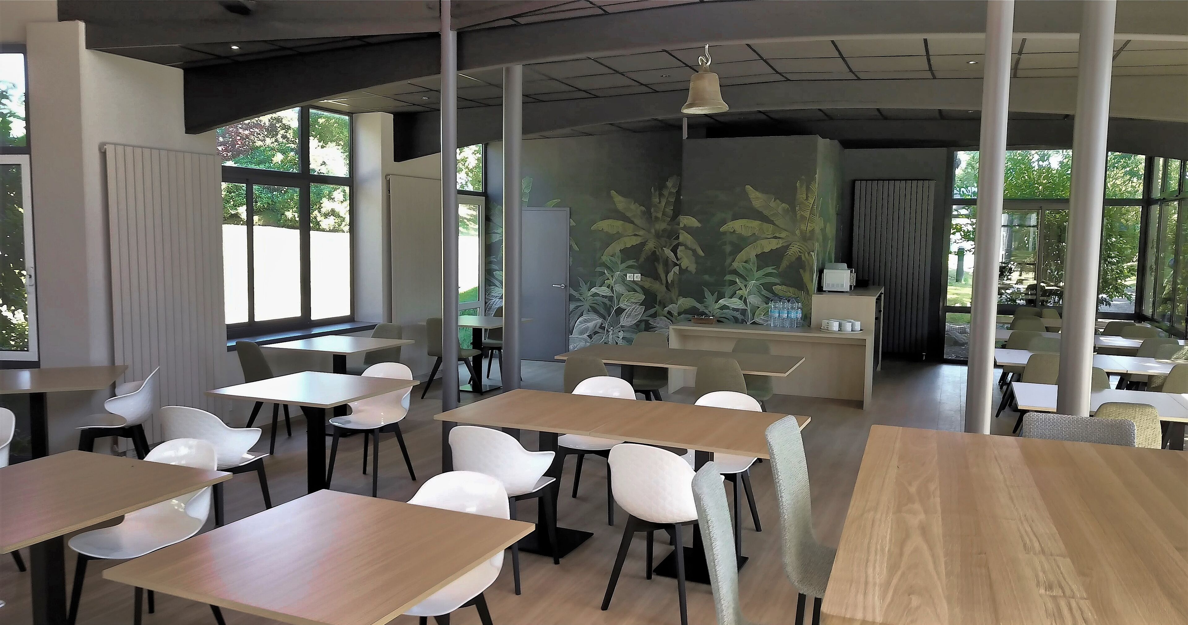 Un comedor moderno con mesas de madera y sillas blancas, ventanas amplias y un mural de plantas tropicales.