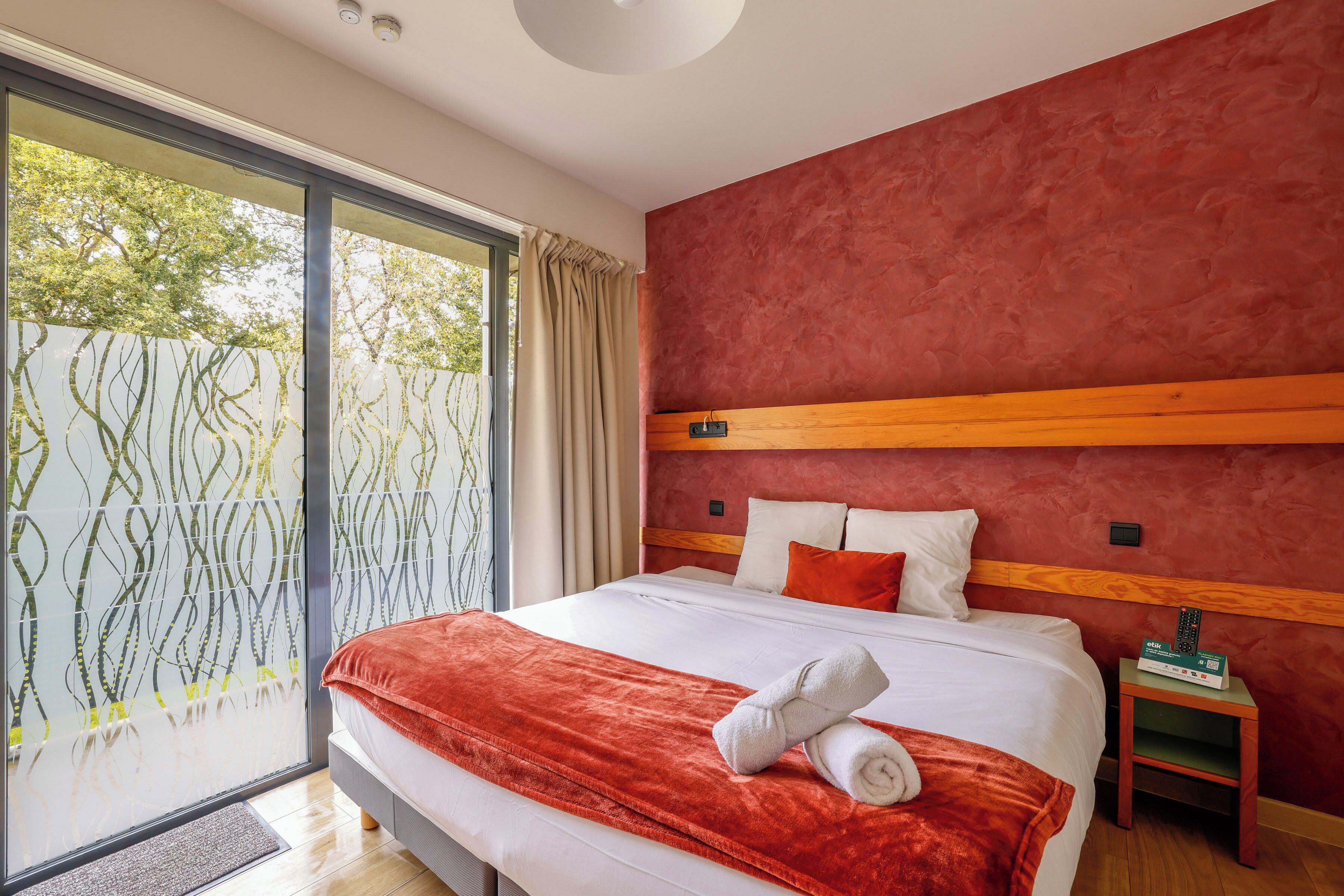 Une chambre d’hôtel moderne avec un grand lit, un mur d’accent rouge et une porte-fenêtre coulissante donnant sur un balcon.