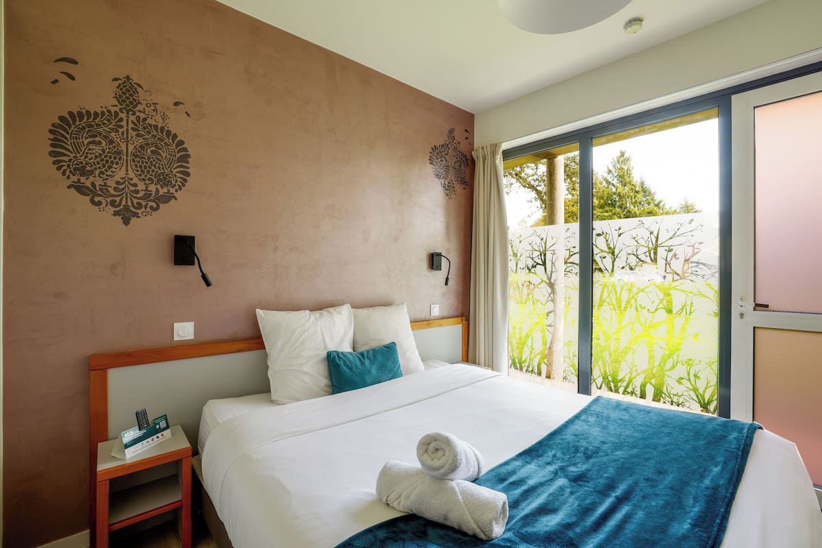 A hotel room with a bed, a nightstand, a wall-mounted lamp, and a view of greenery through a sliding door.