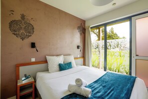 A hotel room with a bed, a nightstand, a wall-mounted lamp, and a view of greenery through a sliding door.