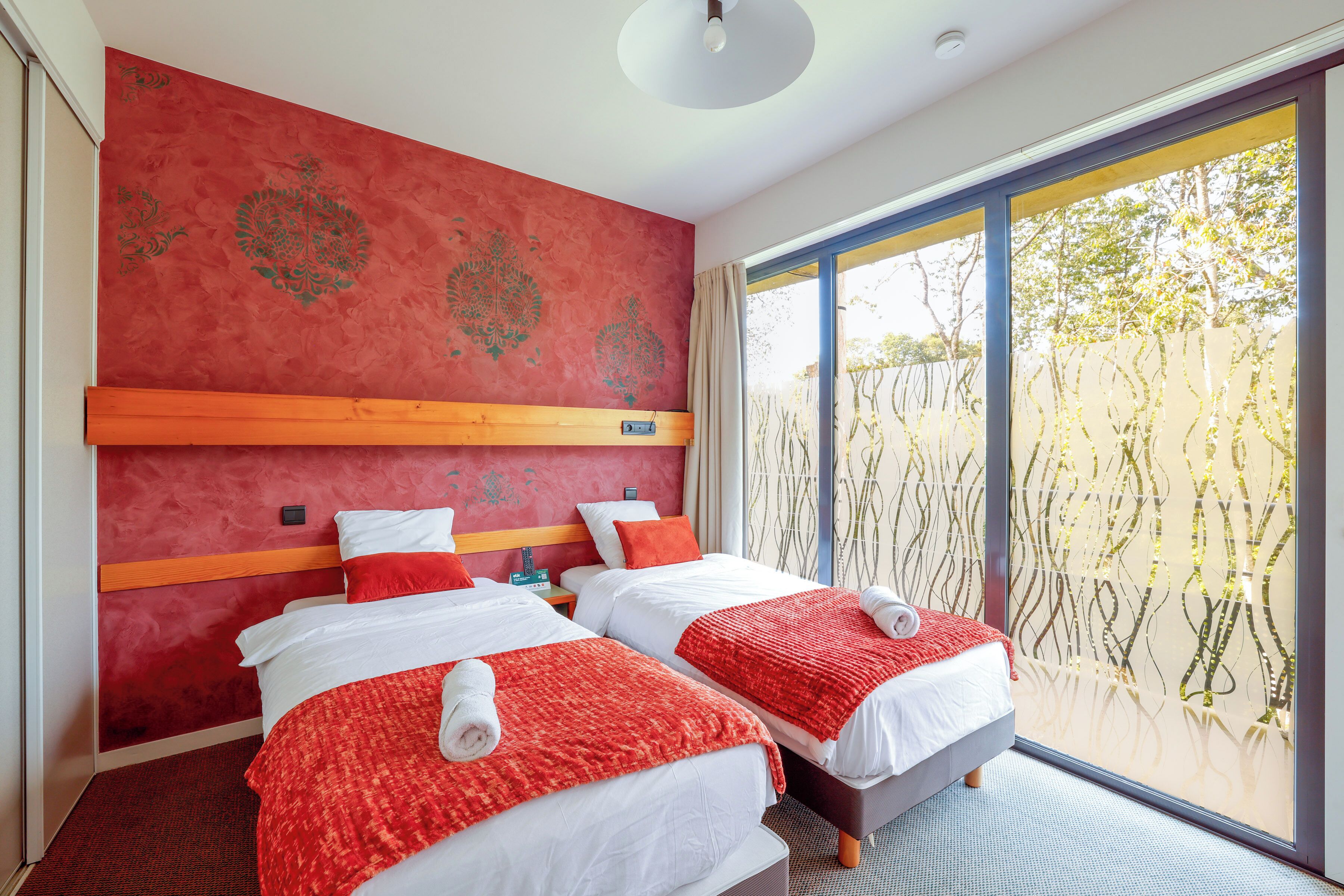 A hotel room with two beds, red and white bedding, a wooden headboard, and large windows with a view of trees.