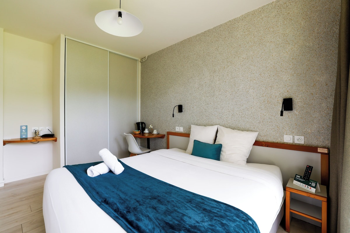 A hotel room with a bed, a desk, a chair, a small table, and a nightstand with a remote control.