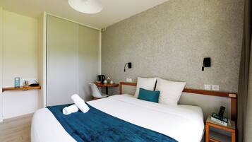 A hotel room with a bed, a desk, a chair, a small table, and a nightstand with a remote control.