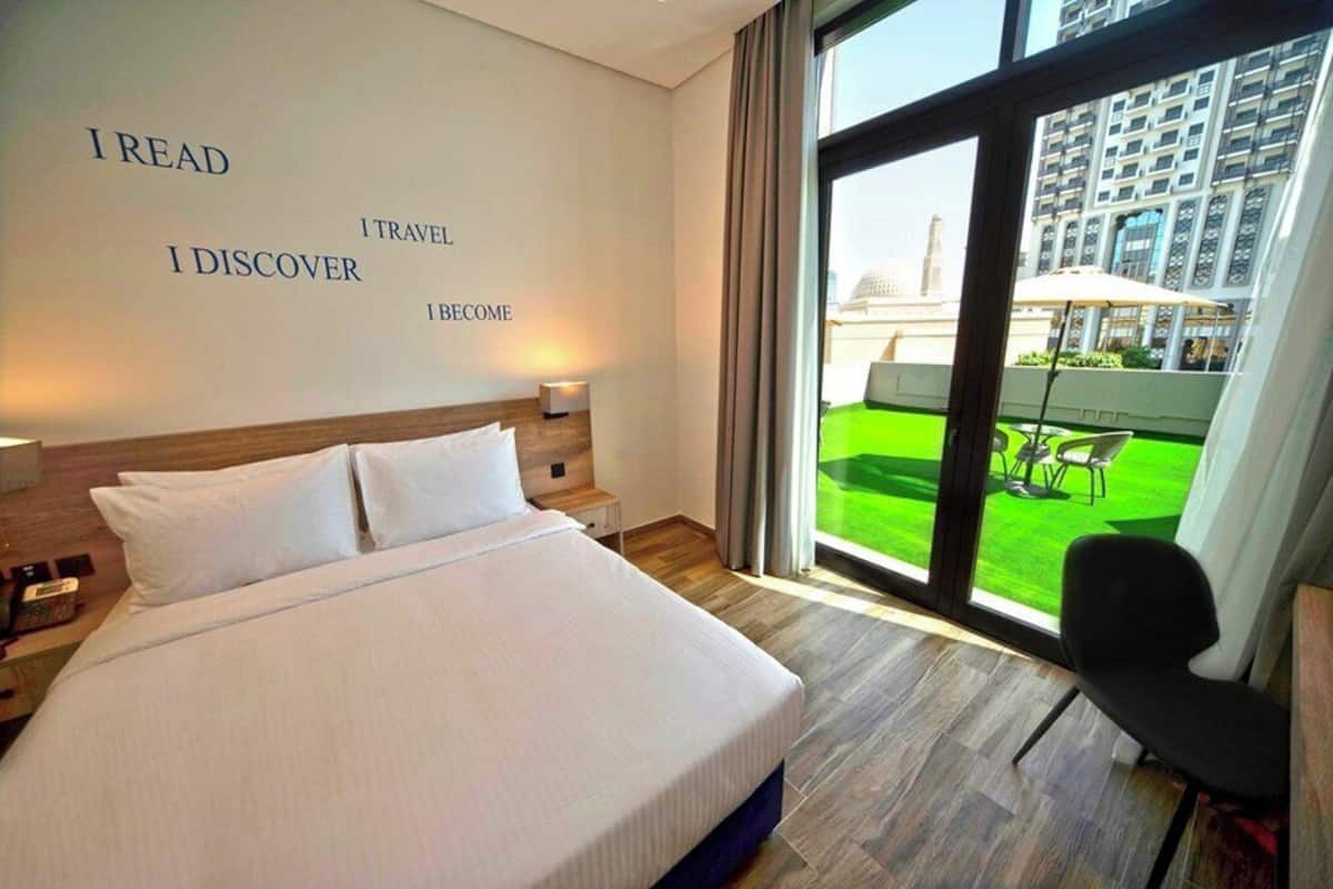 Camera d'albergo con un letto grande, un comodino, una parete con frasi motivazionali e vista su una zona verde attraverso un'ampia finestra.