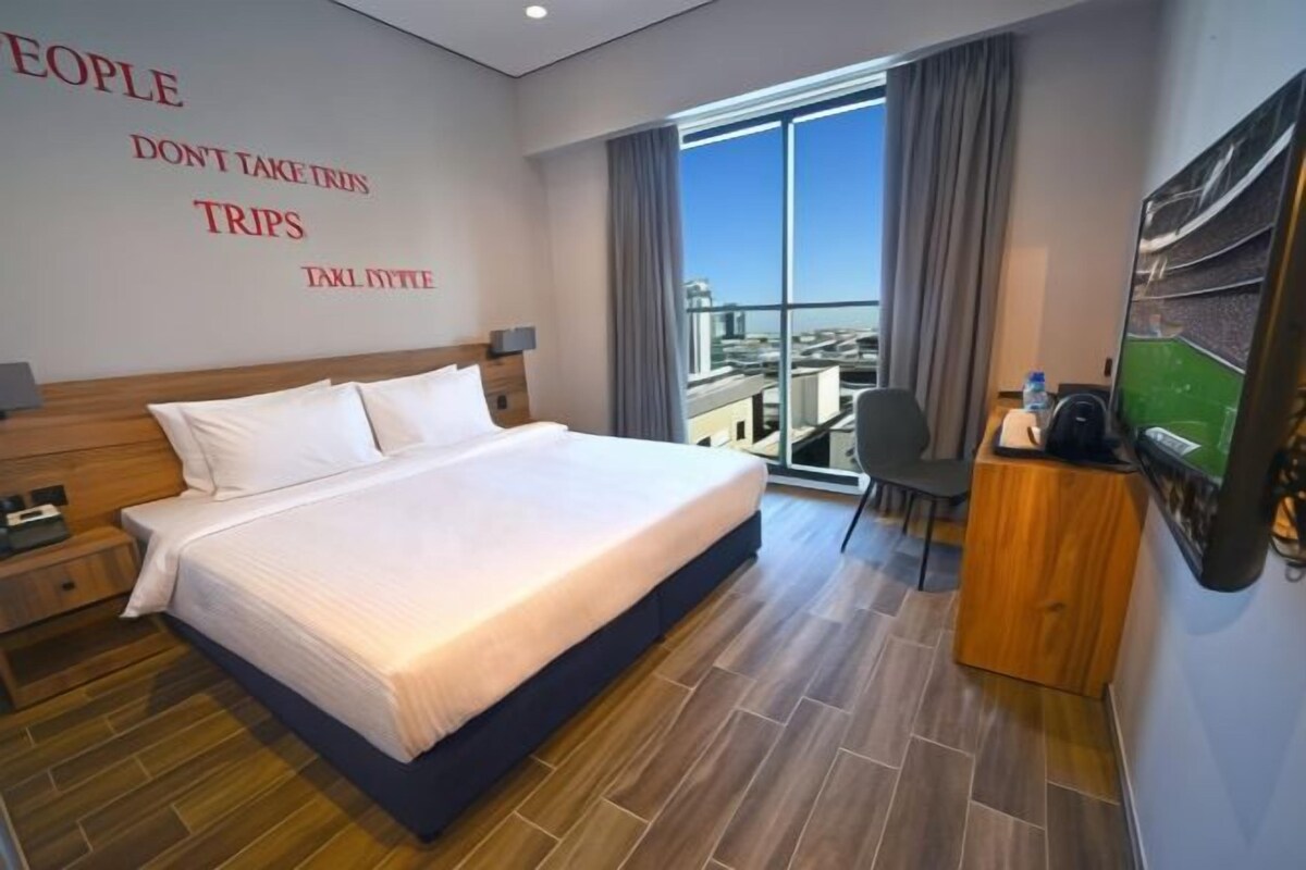 Quarto de hotel com uma cama grande, uma escrivaninha com cadeira, televisão e vista para a cidade pela janela.