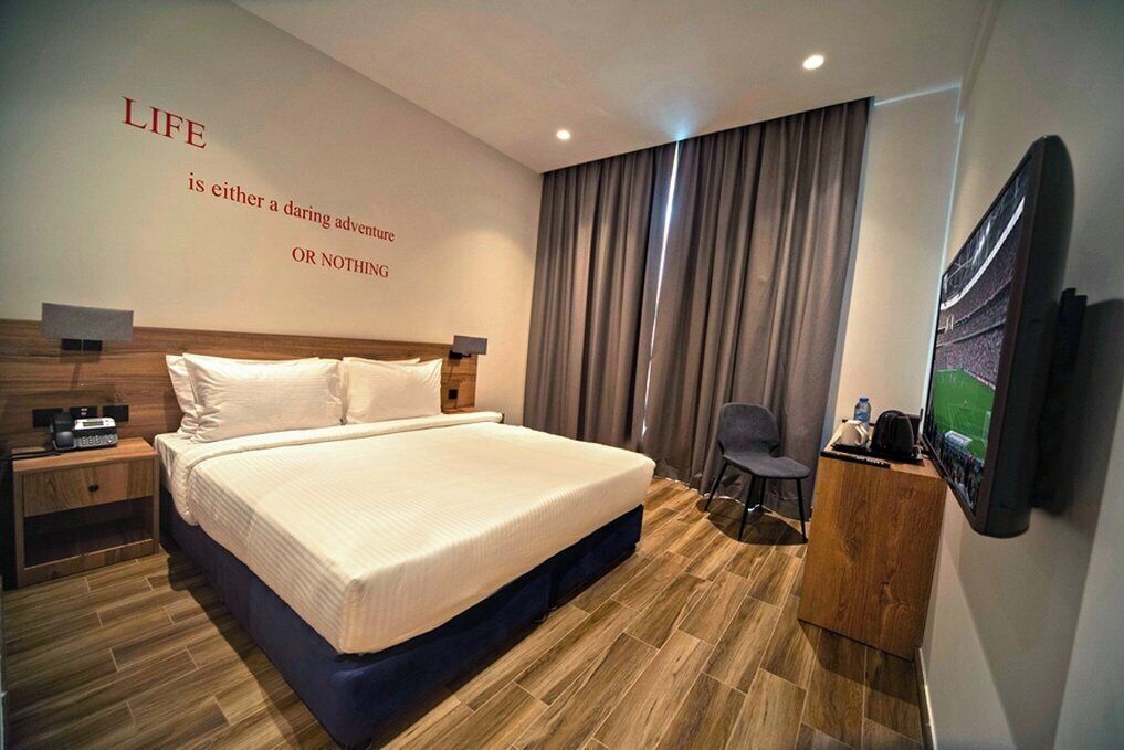 A hotel room with a large bed, a desk with a chair, a TV, and a wall with a motivational quote.