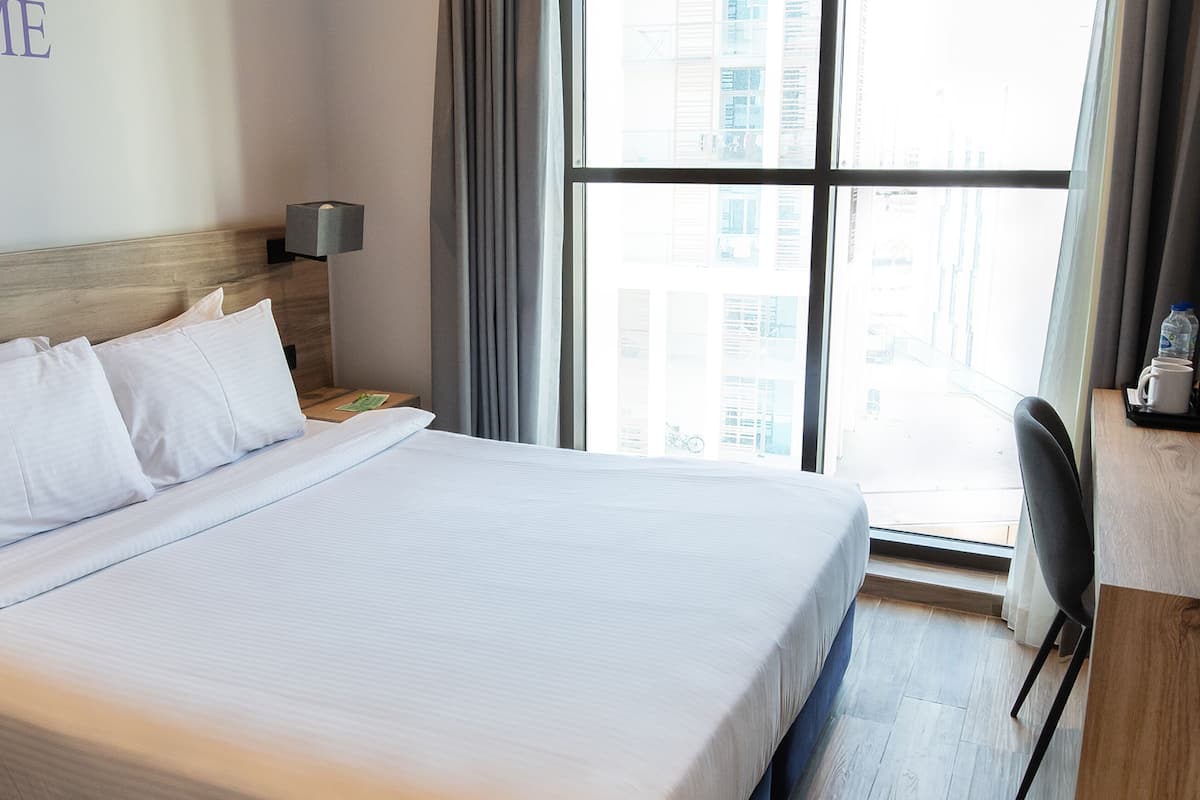 Camera d'albergo con un letto grande, un comodino, una lampada, una sedia e vista sulla città dalla finestra.