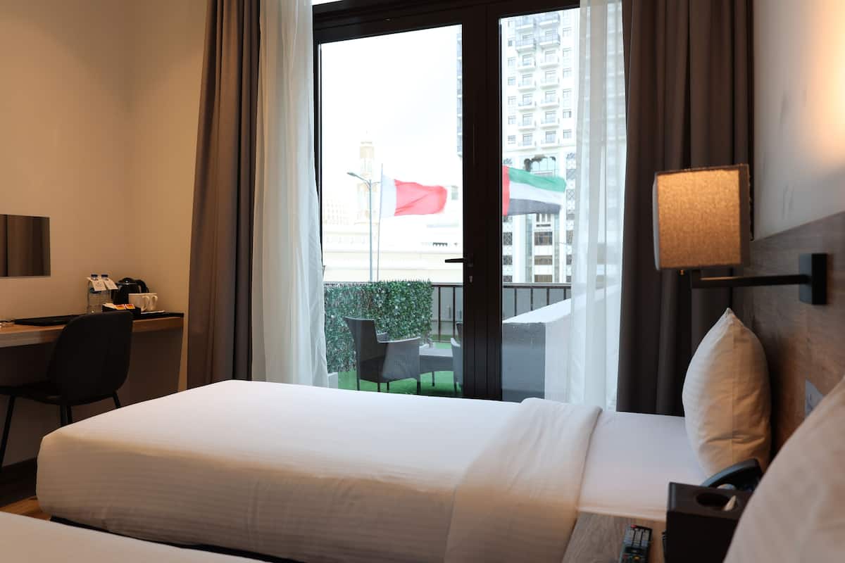 Una camera d'albergo con un letto, una scrivania con una sedia, una lampada e vista sulla città da una grande finestra.