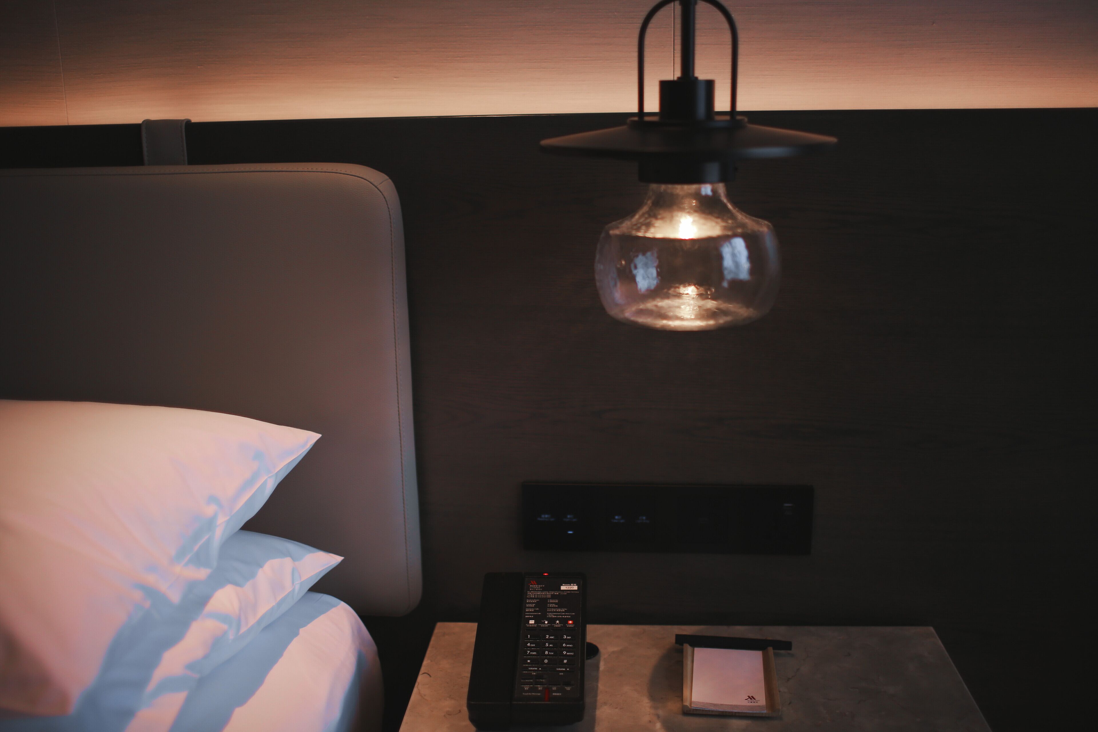 A hotel room with a bed, a bedside table with a remote control, a lamp, and a wall-mounted control panel.