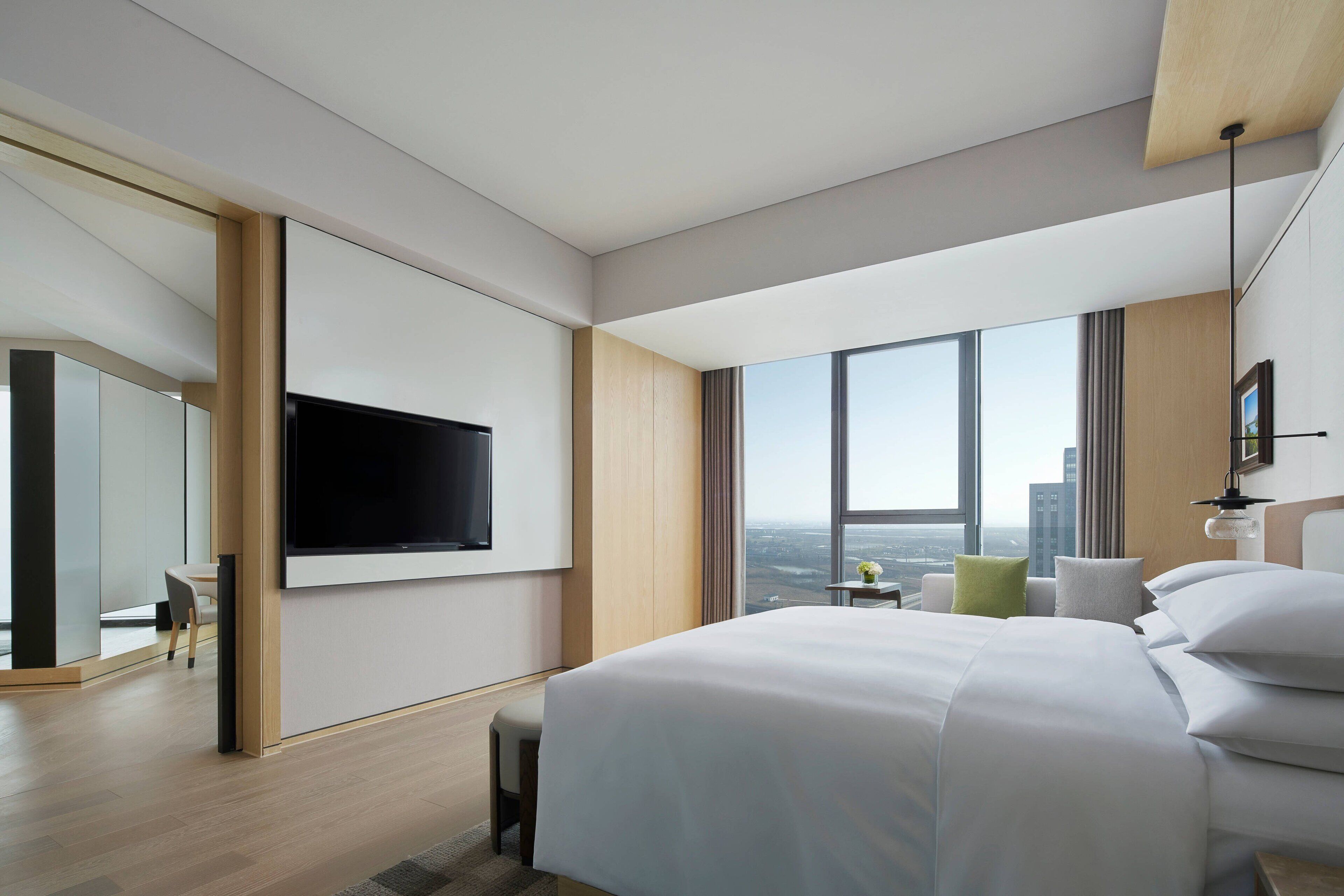 A modern hotel room with a large bed, a flat-screen TV mounted on the wall, a small table with a vase, and a view of the city through large windows.