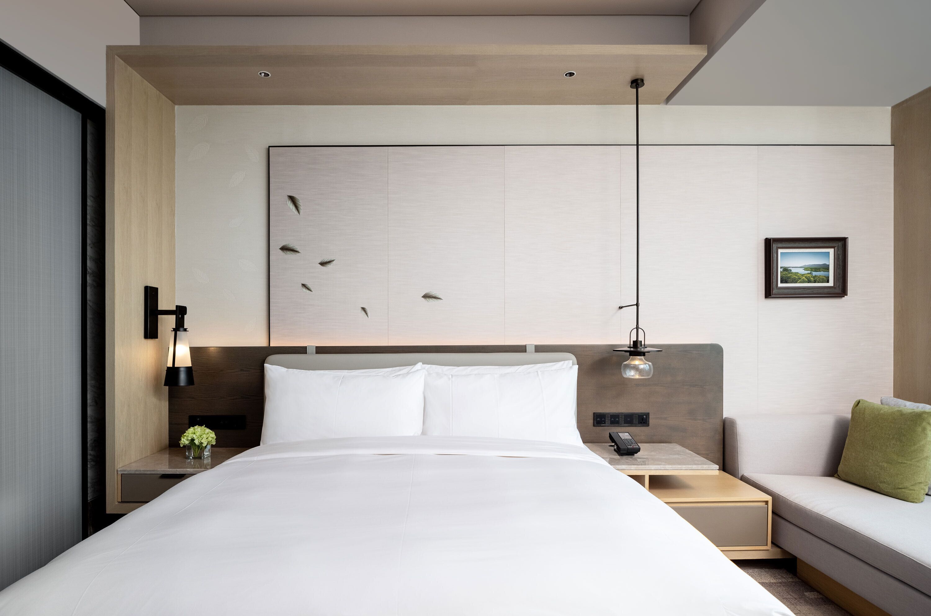 Habitación de hotel moderna con una cama grande, una mesita de noche con lámpara, una pequeña mesa con teléfono, un sofá con almohada y un cuadro colgado en la pared.