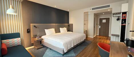 A modern hotel room with a large bed, a desk, a chair, and a sofa.