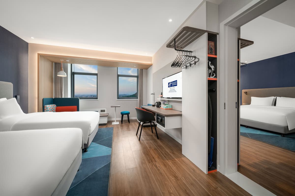 A modern hotel room with two beds, a desk, and a view of the city.