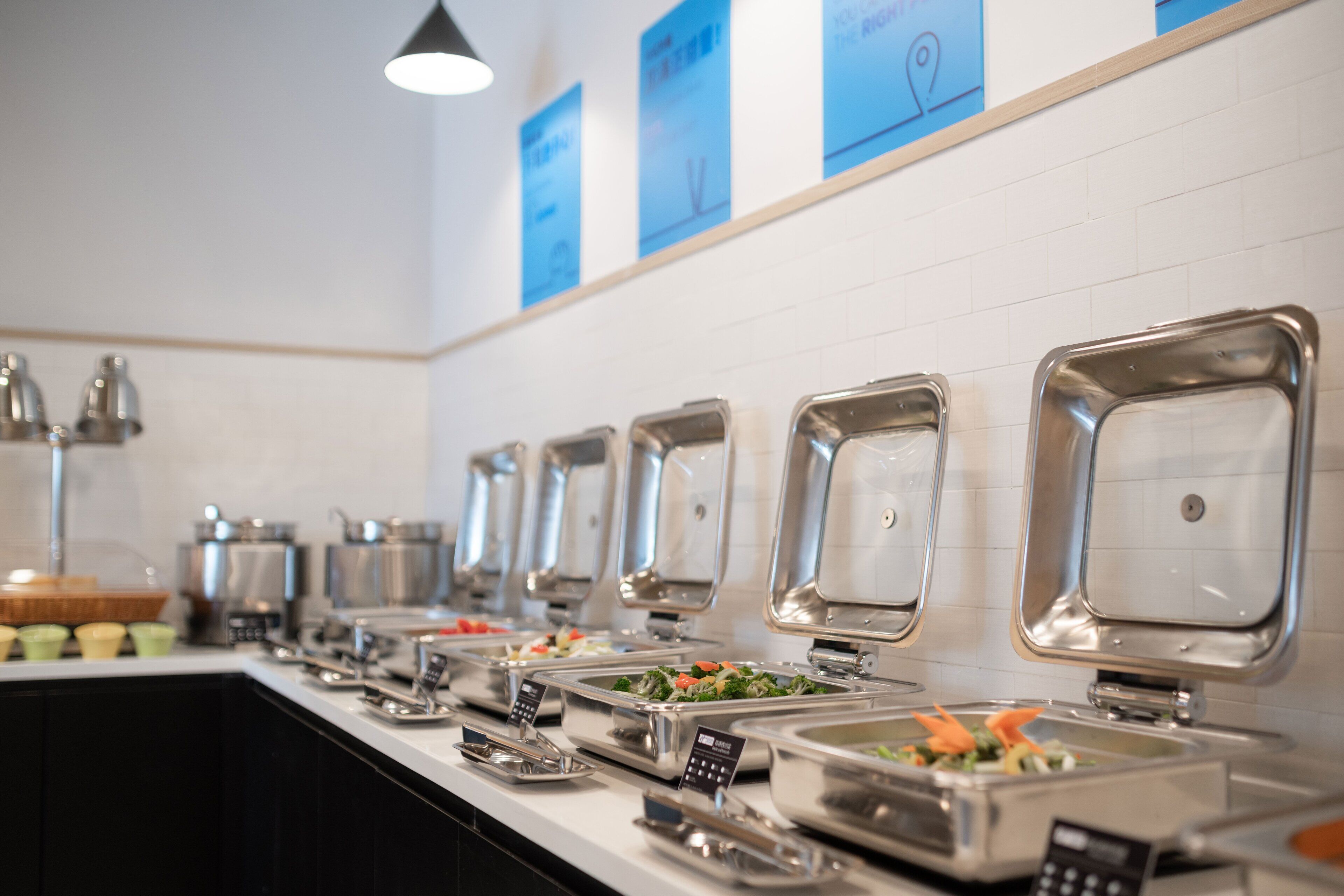 A buffet line with multiple self-service food stations, each with a metal tray and a small sign above.