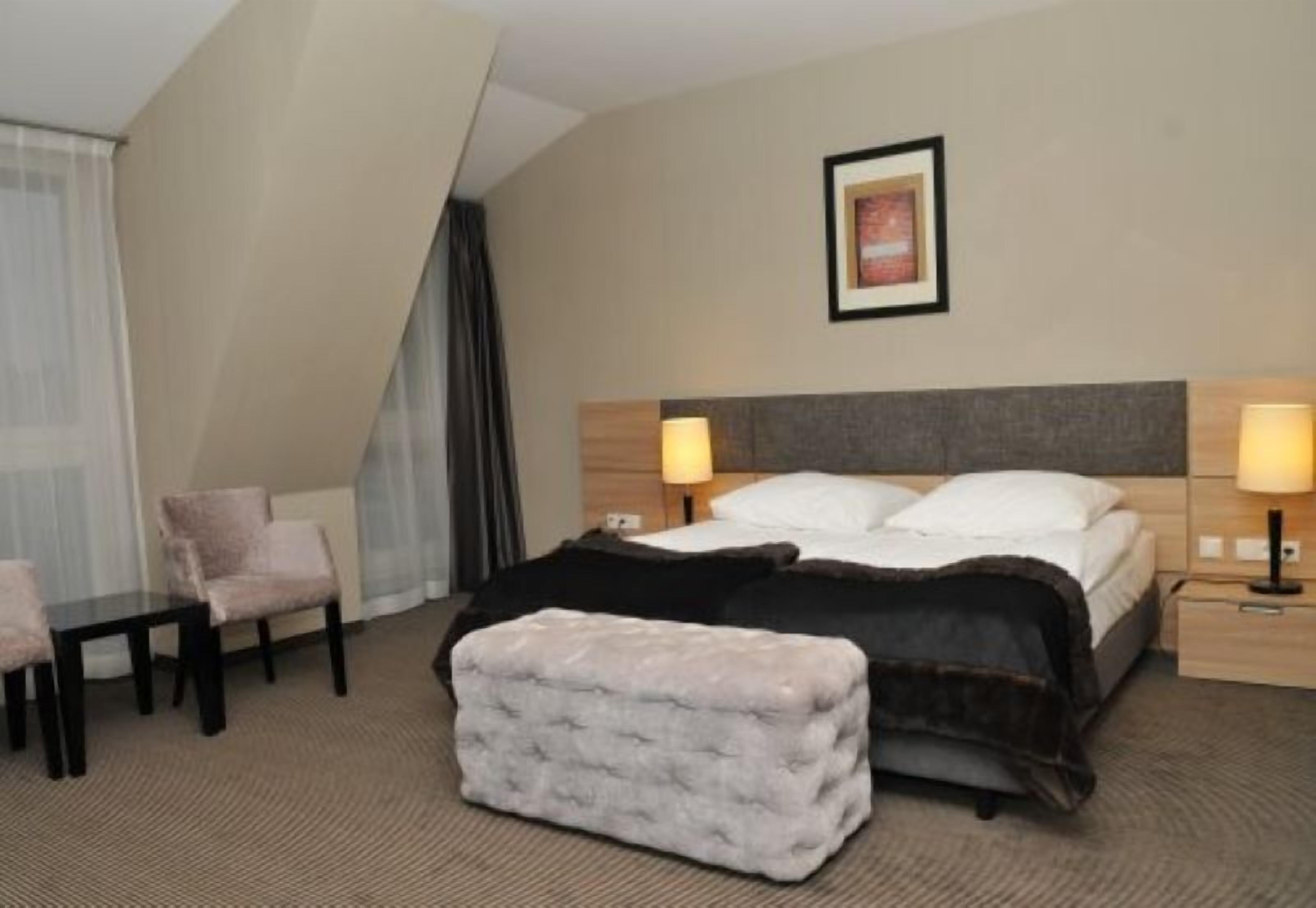 Une chambre d’hôtel avec un lit, deux tables de chevet, une chaise, un petit pouf et un tableau encadré au mur.