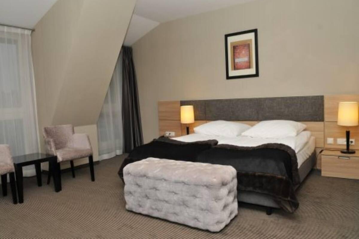 A hotel room with a bed, two bedside tables, a chair, a small ottoman, and a framed picture on the wall.