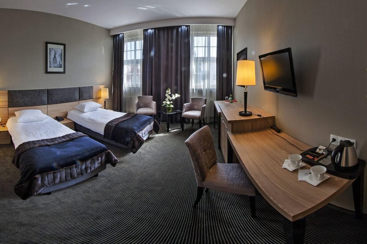A hotel room with two beds, a desk, a chair, a TV, and a window with curtains.